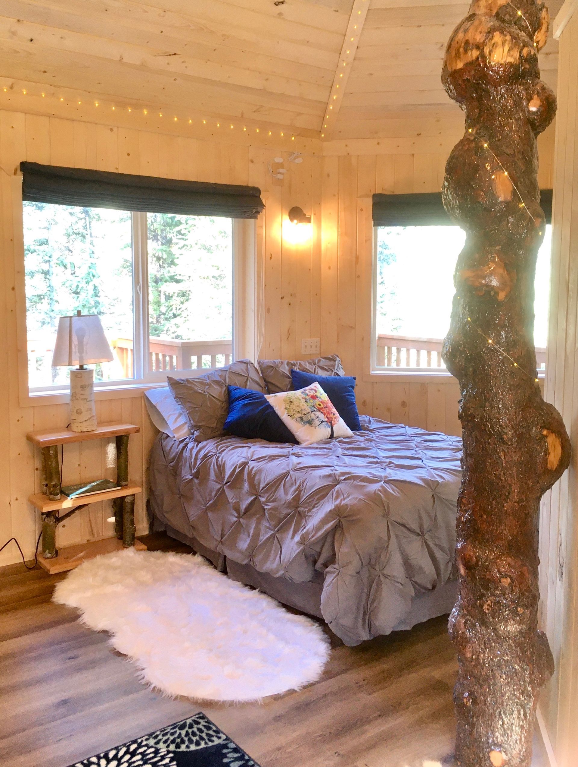 Bedroom in a treehouse with a bed, windows, rug, and a large tree trunk inside.