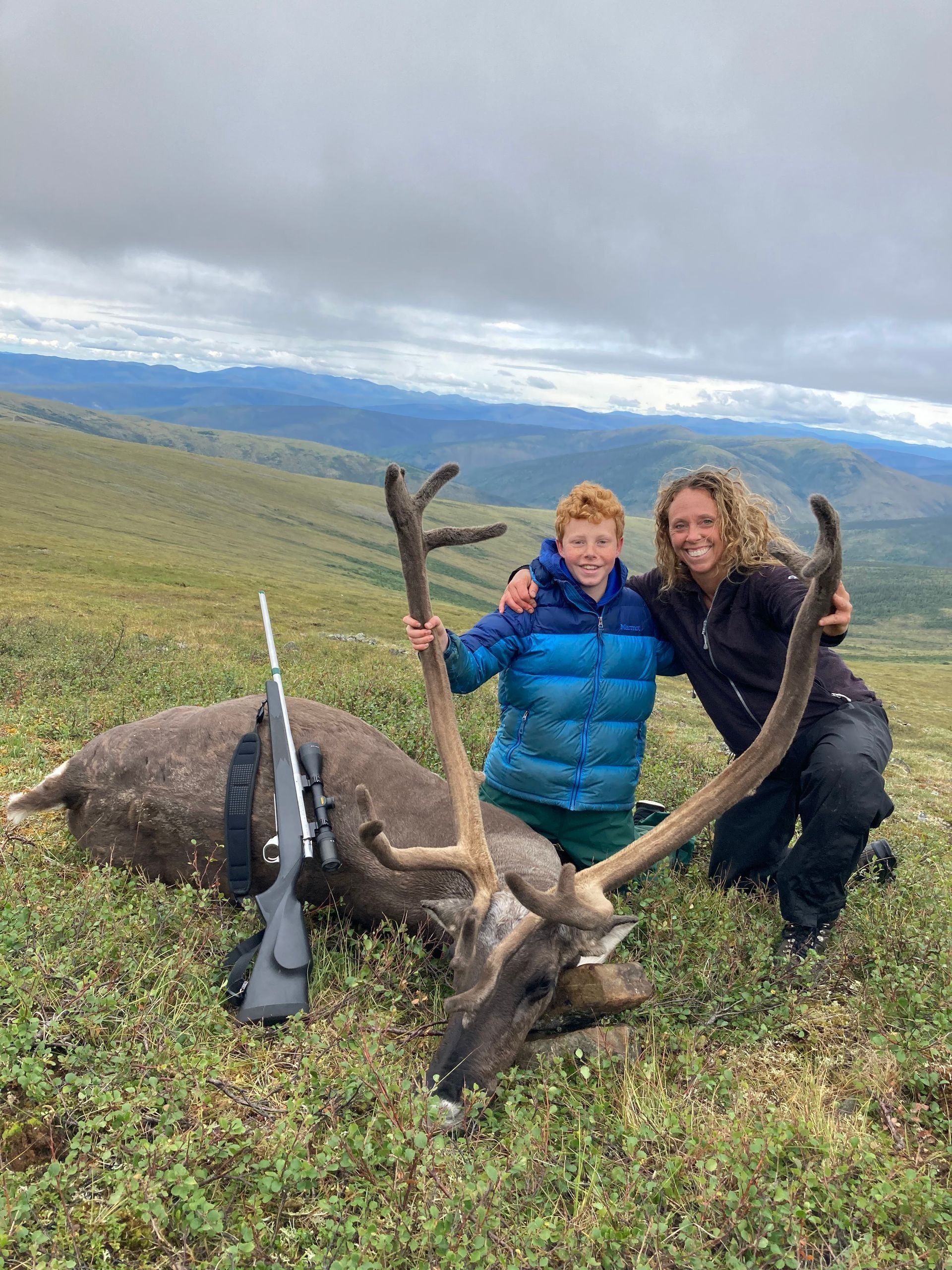 A person and child pose with a large caribou they hunted; rifle nearby, mountains in the background.