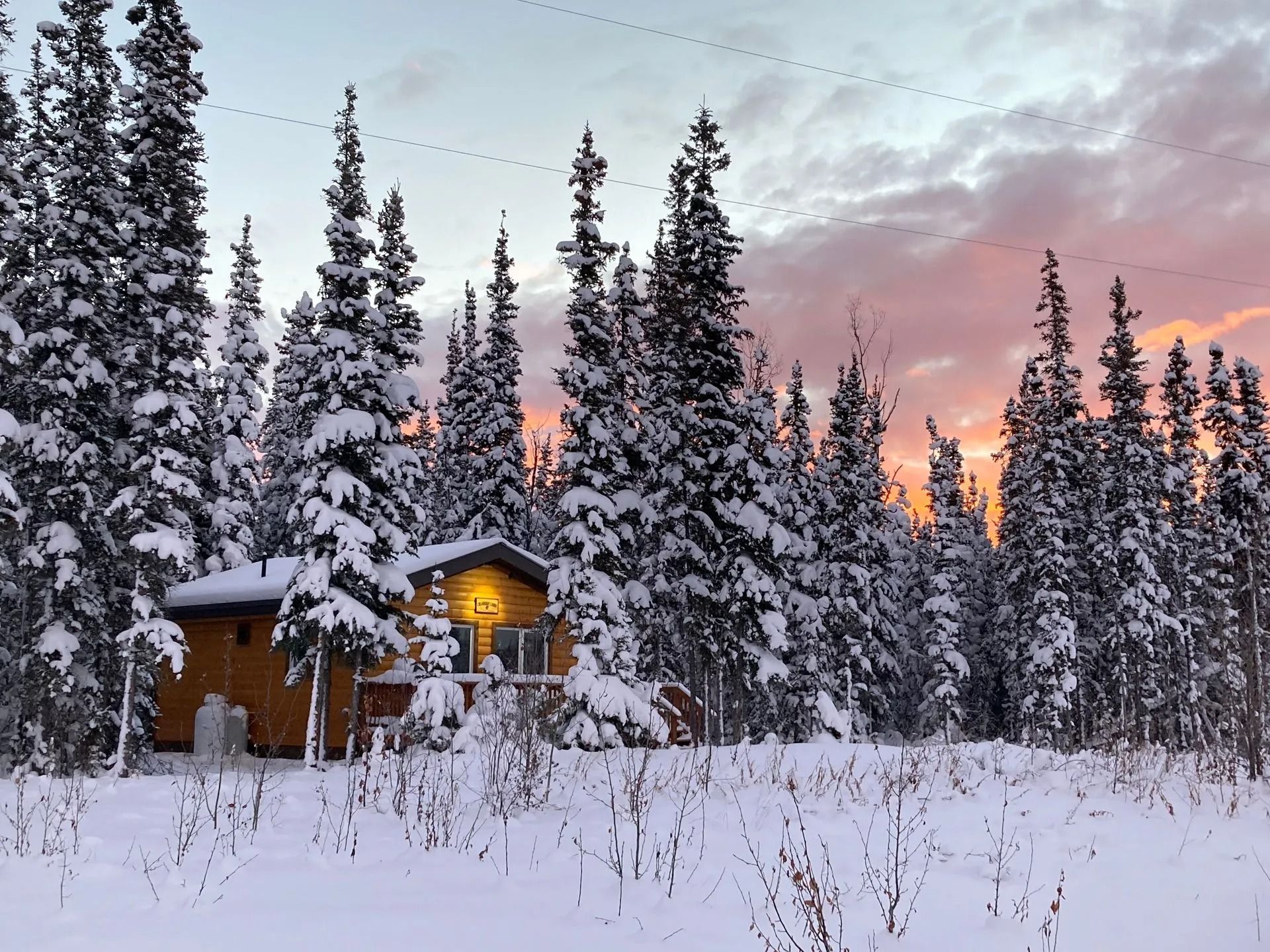 Snow-covered cabin in a forest with an orange and pink sunset in the background.
