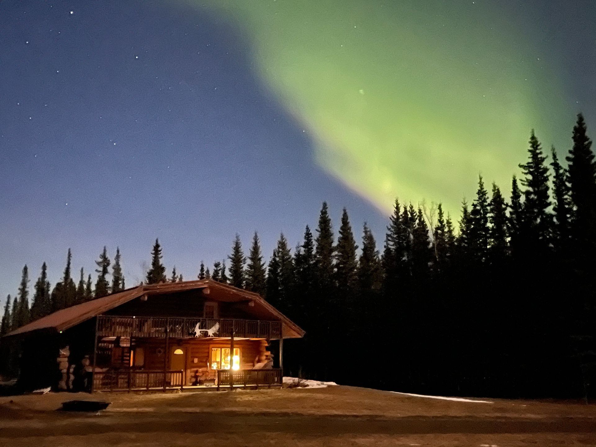 Cabin illuminated with lights under aurora borealis in green and blue night sky. Forest in the background.