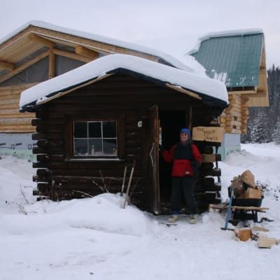 A man in a red jacket stands in front of a snow covered log cabin