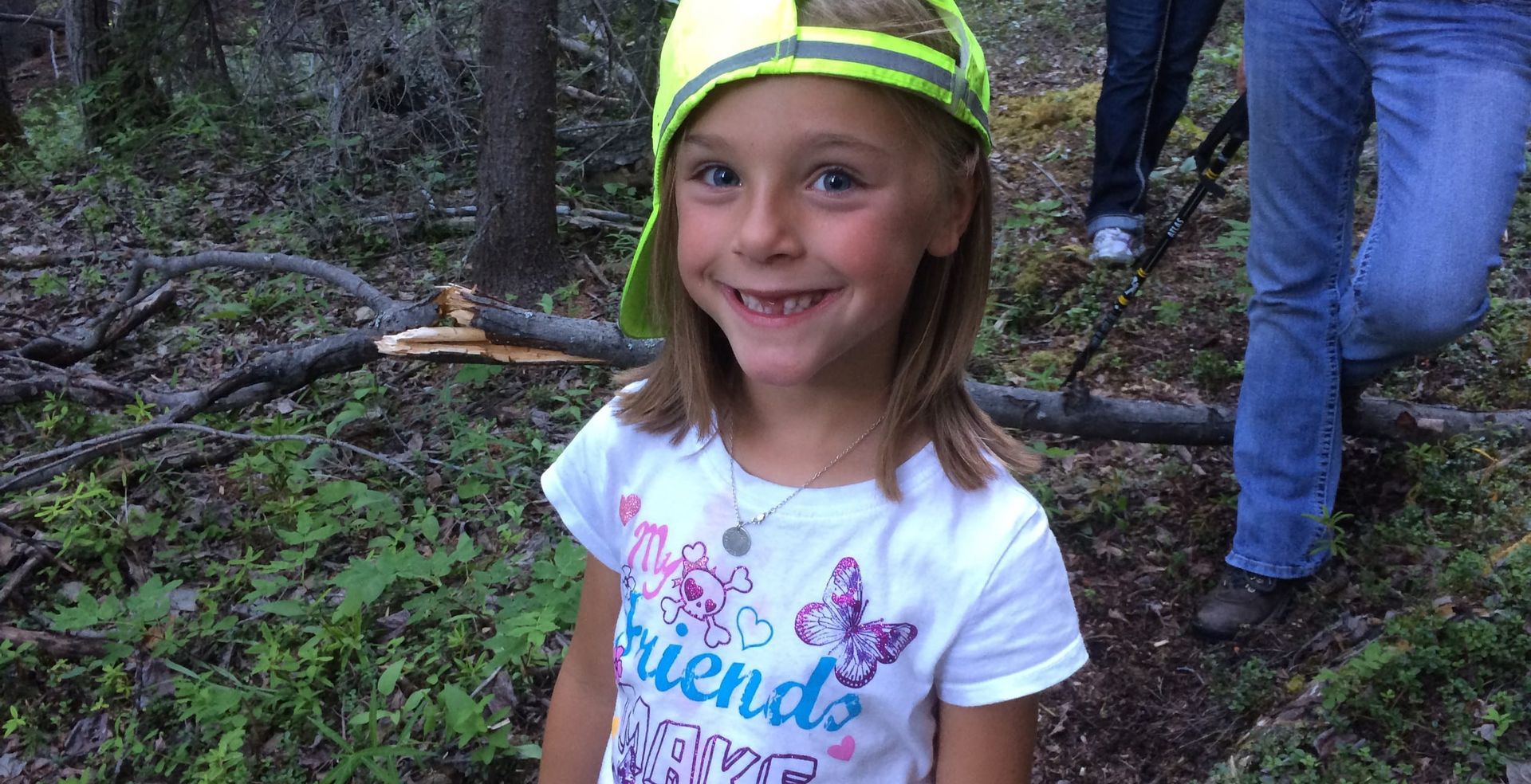 A little girl wearing a yellow hat and a white shirt is standing in the woods.