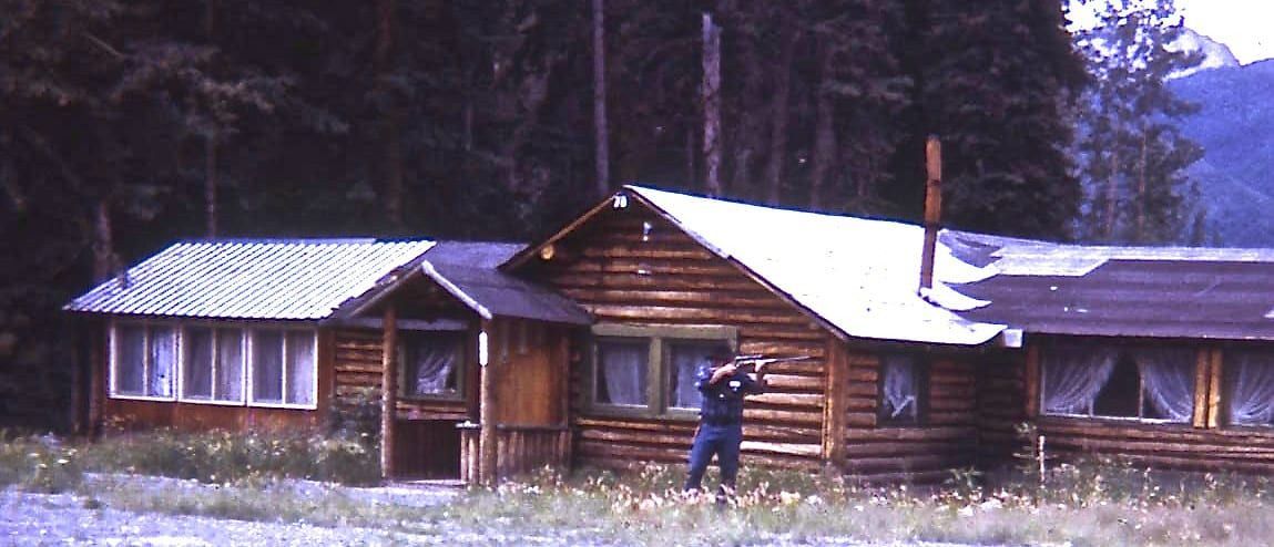 A man stands in front of a log cabin in the woods