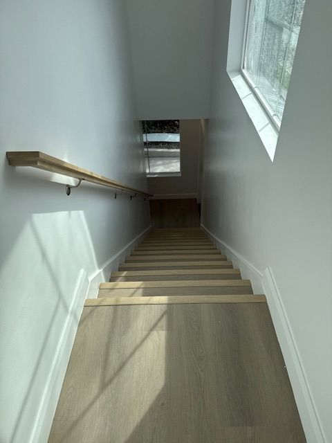 Staircase leading downwards with a wooden handrail on the left wall and a window on the right, both with white walls.