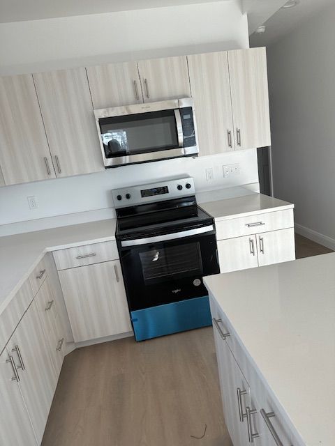 Modern kitchen with light wood-grain cabinets, white countertops, and stainless steel appliances.
