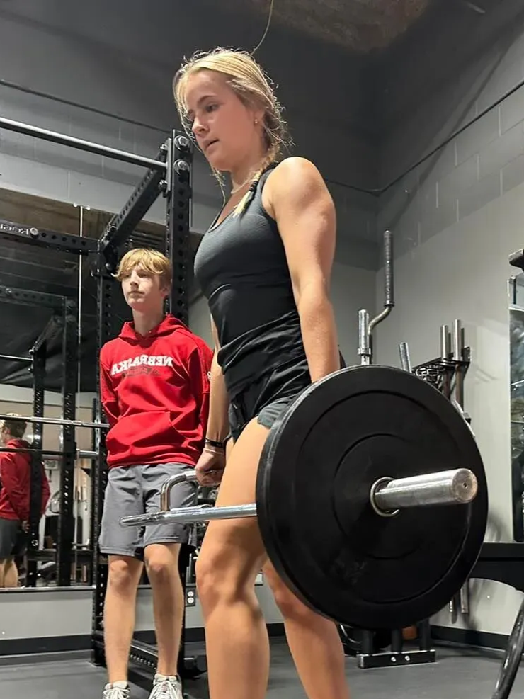 Woman deadlifts in a gym, supervised by a young man. She wears black workout clothes. Gray and black gym setting.
