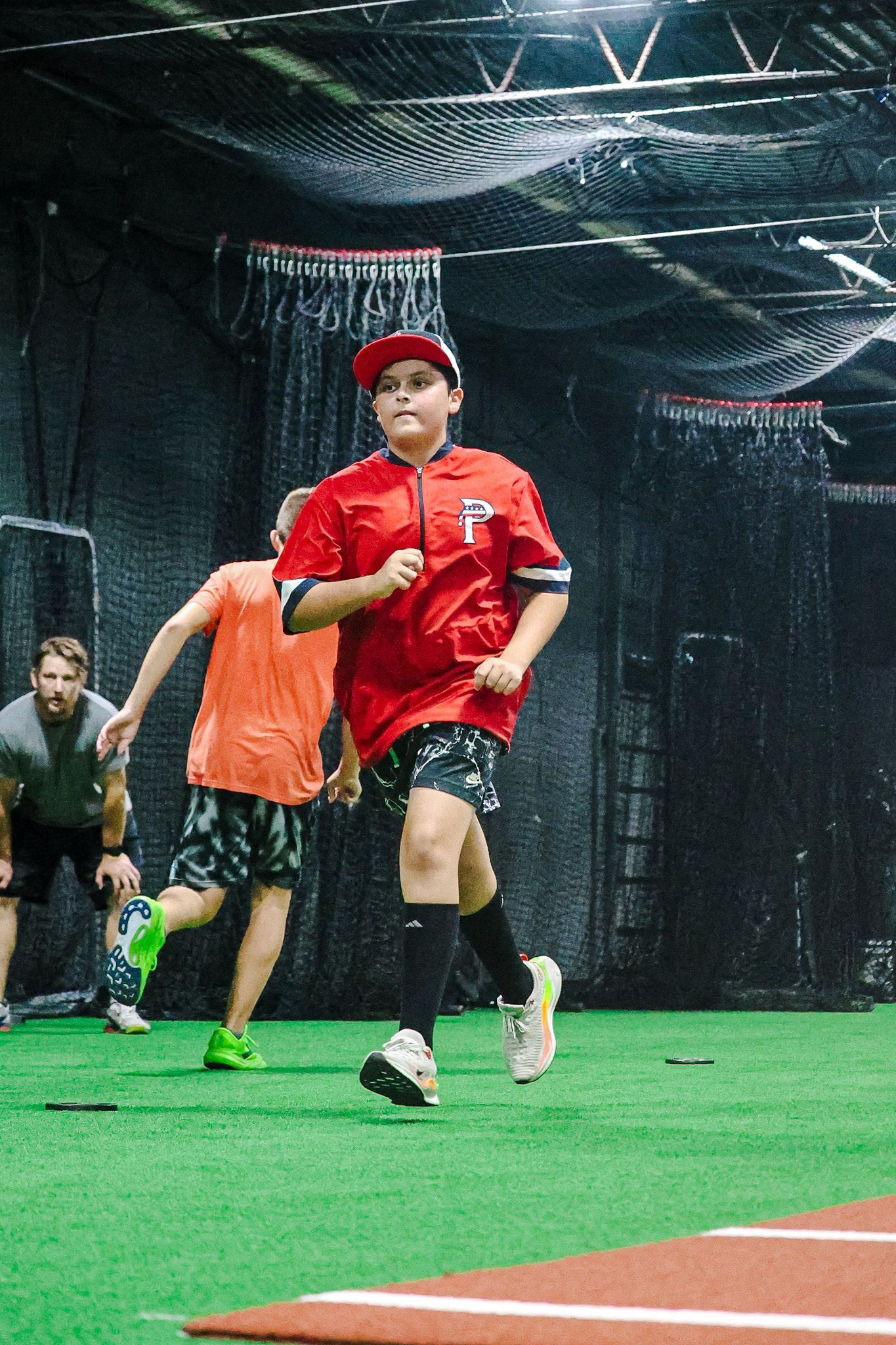Boy in red jersey running toward camera on a baseball field, other boys running behind, and a coach looking on.