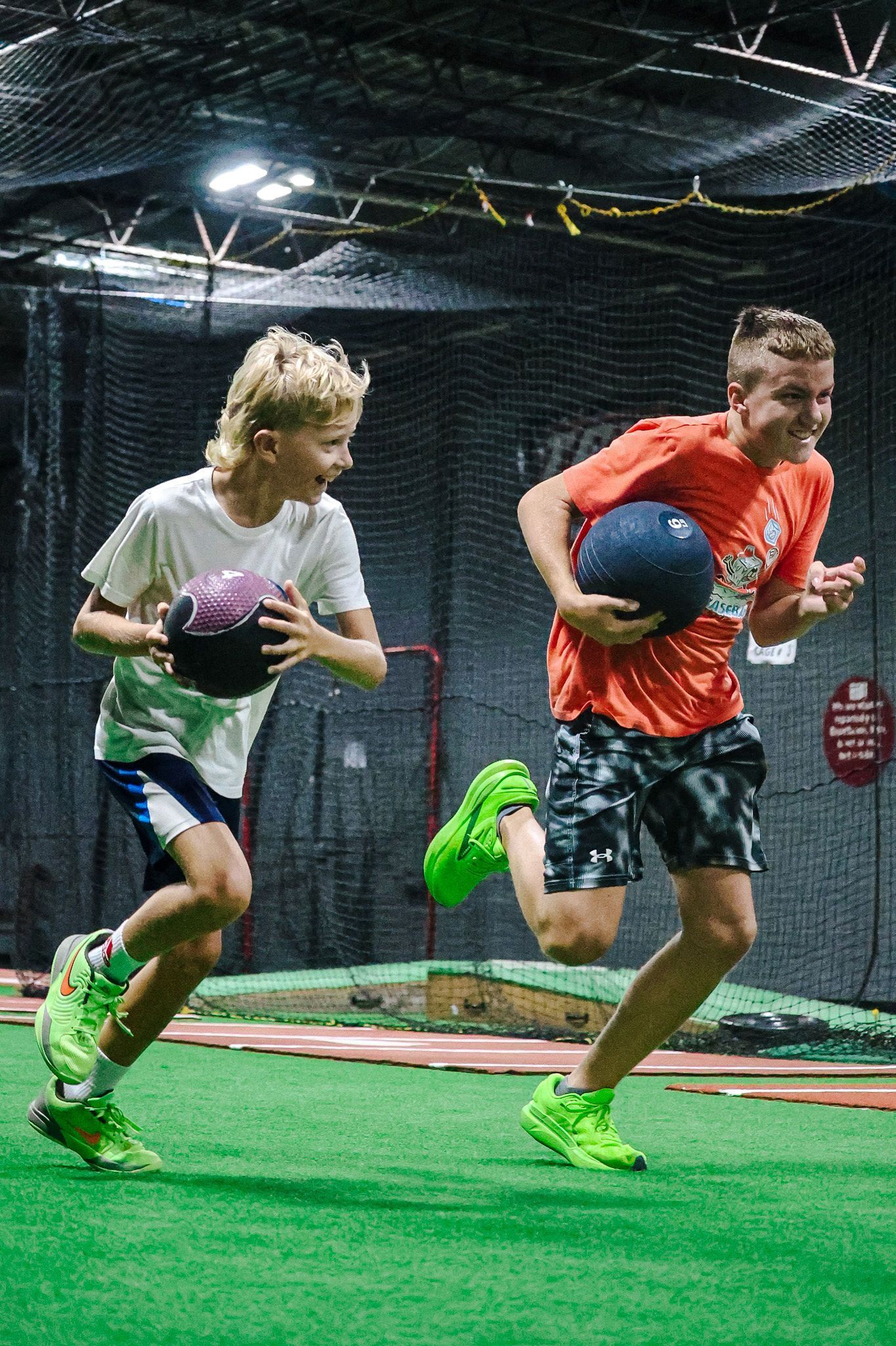 Two boys running indoors, holding medicine balls, one in white, the other in orange, on a green turf field.