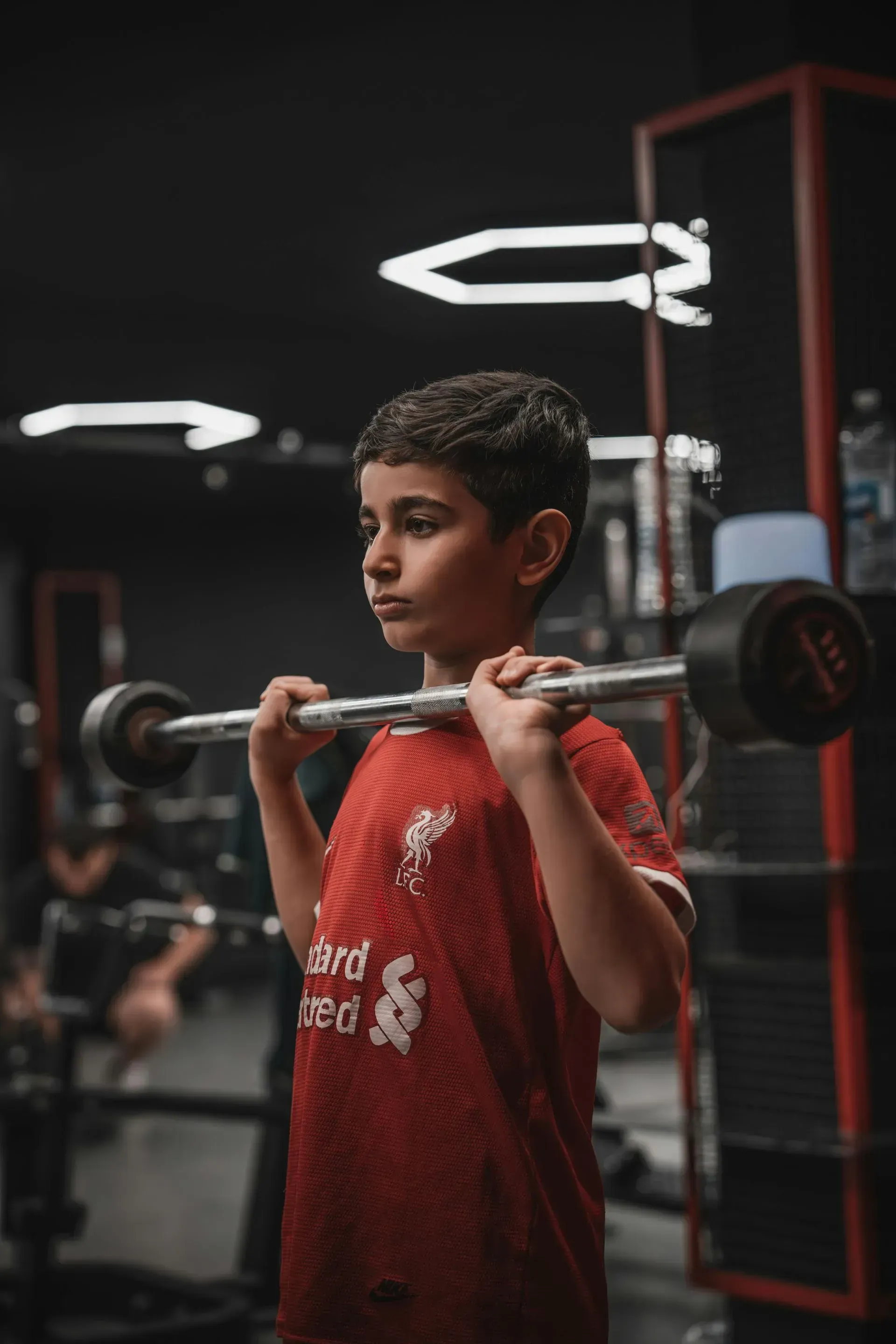 Boy in red jersey holding a barbell on his shoulders at a gym, with a determined expression.