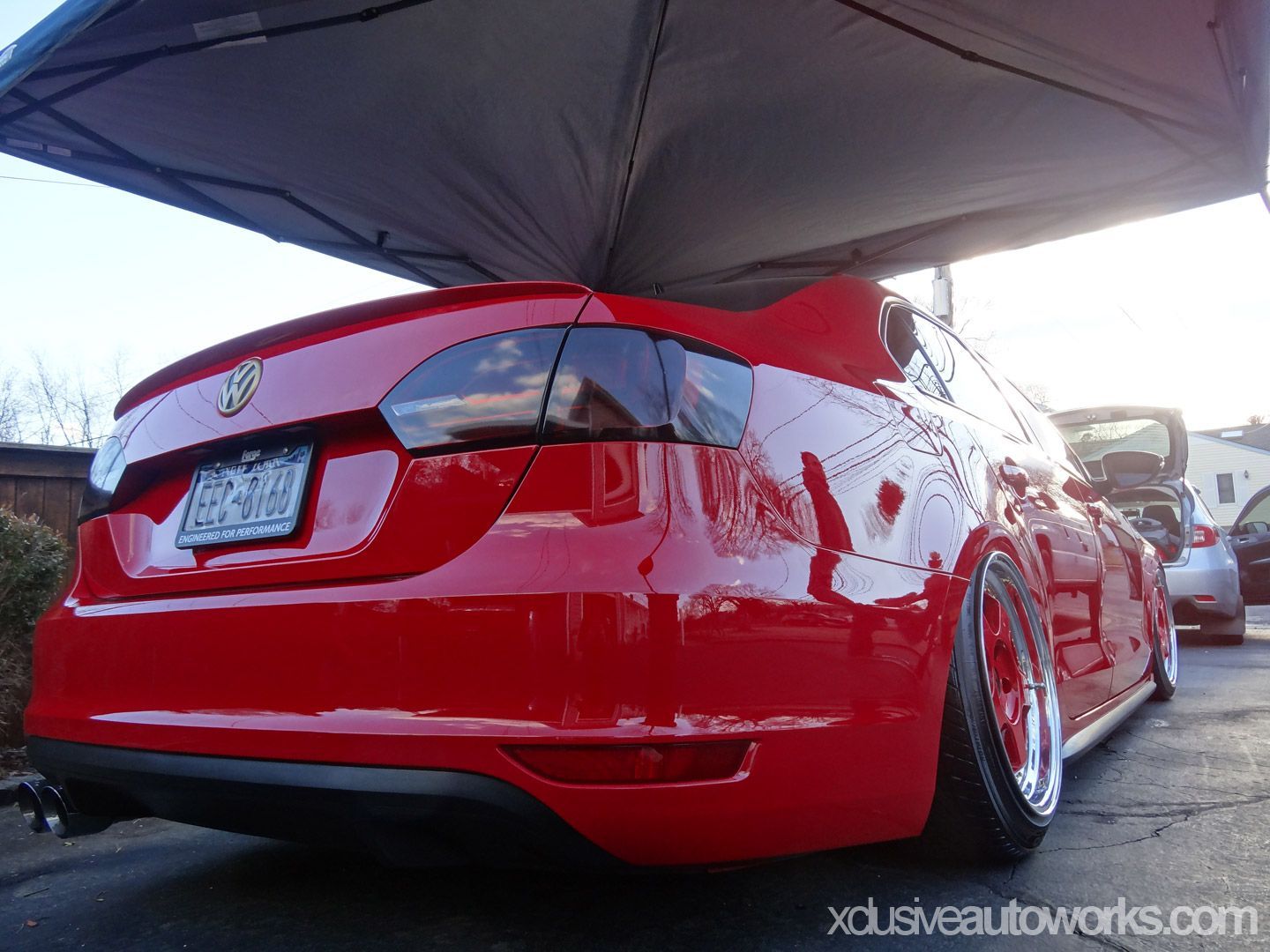 A red car is parked under a tent with xclusiveautoworks.com written on the bottom