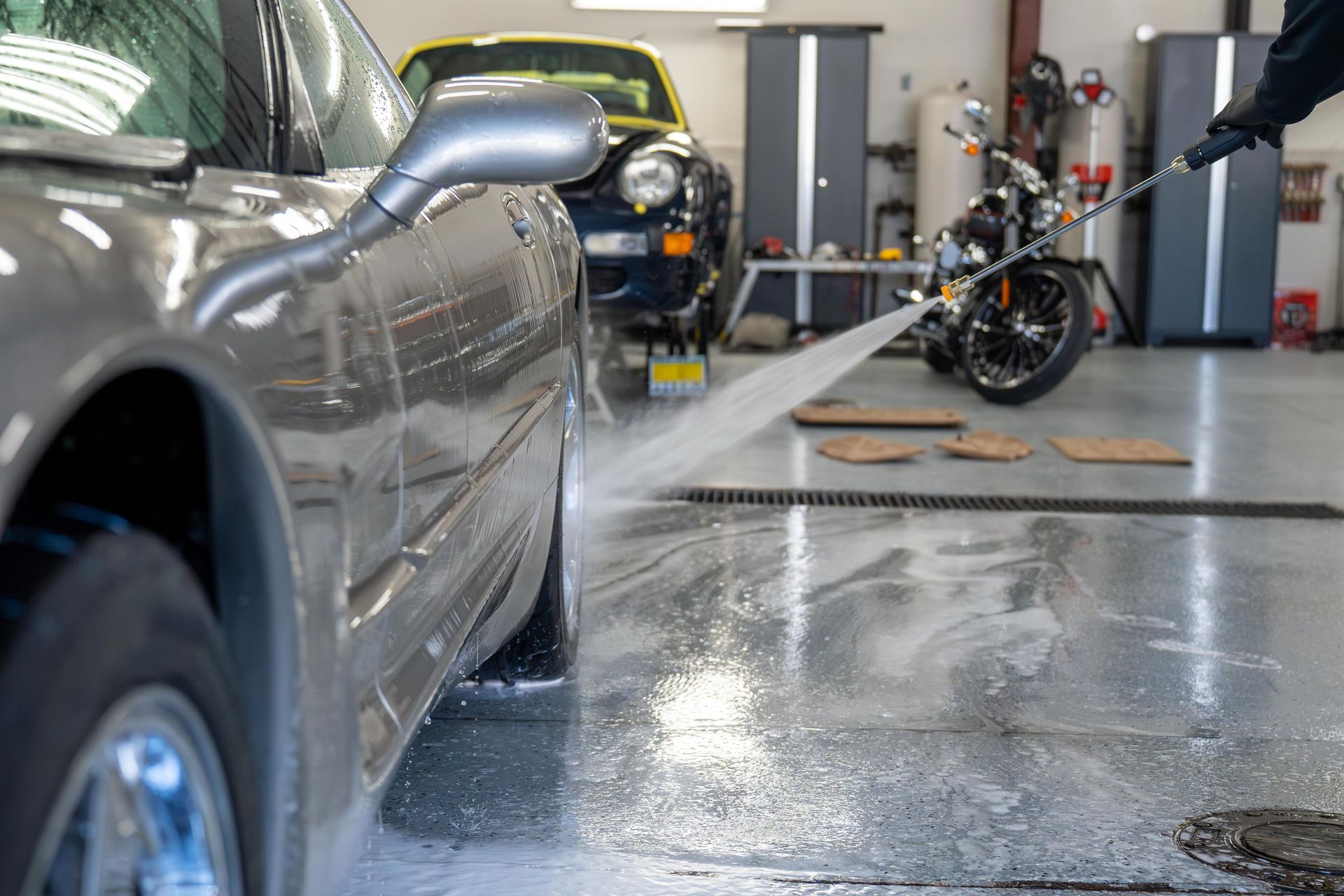 A person power-washes the wheel of a gray car inside a garage, with a motorcycle parked in the background.