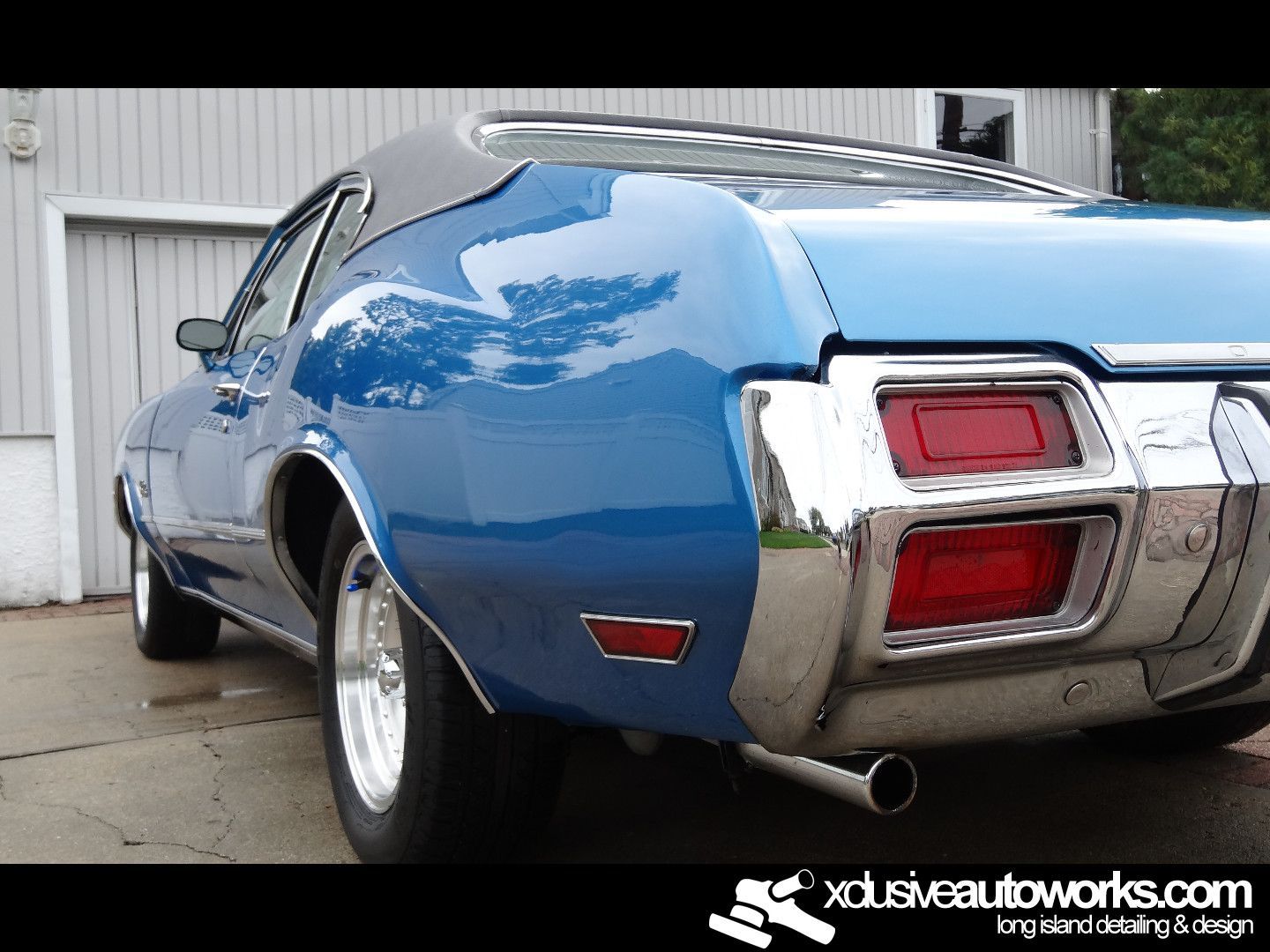 A blue car is parked in front of a garage with xdusveautoworks.com written on the bottom
