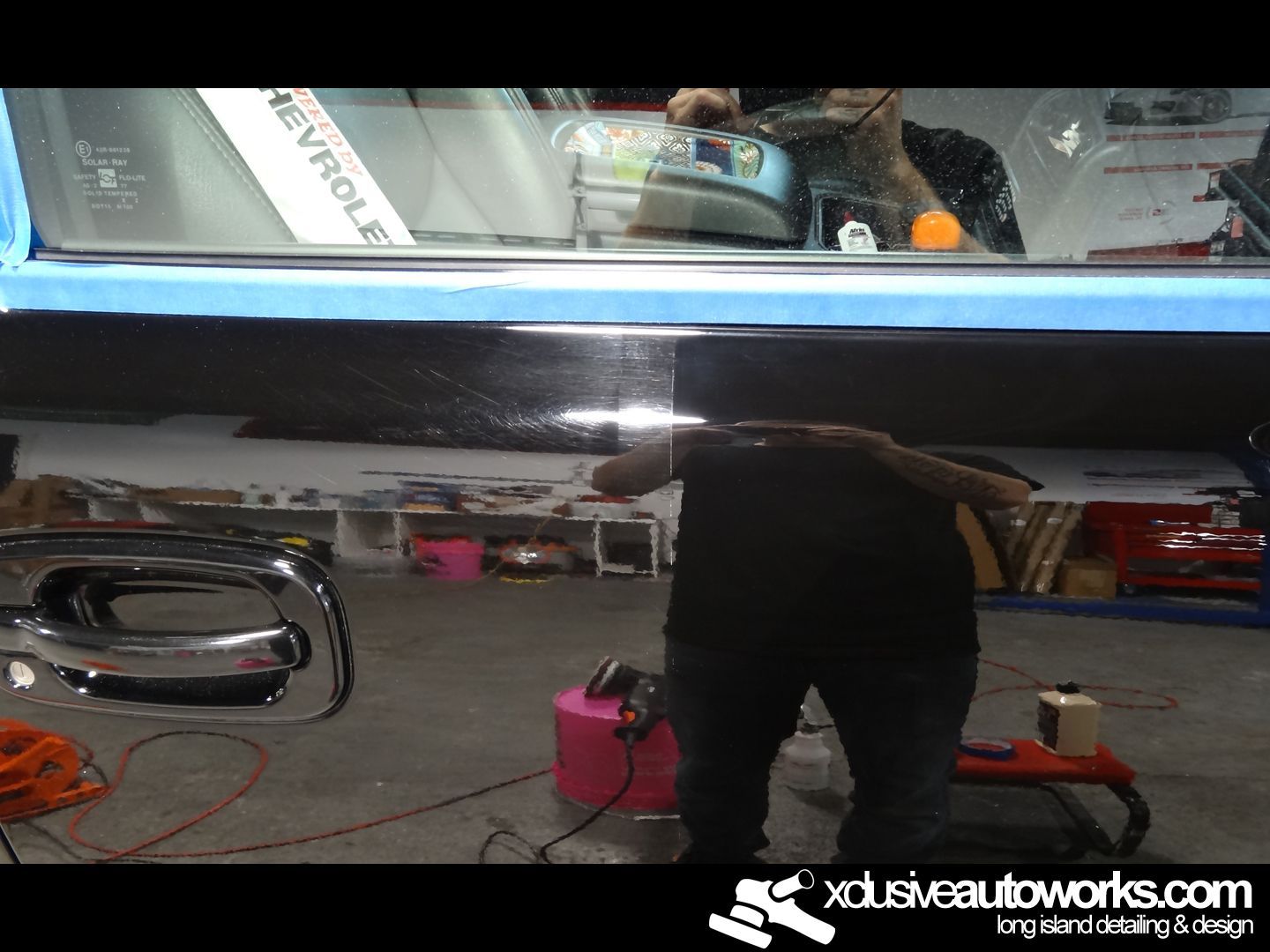 A man is polishing a car in a garage with the website xdusveautoworks.com in the corner