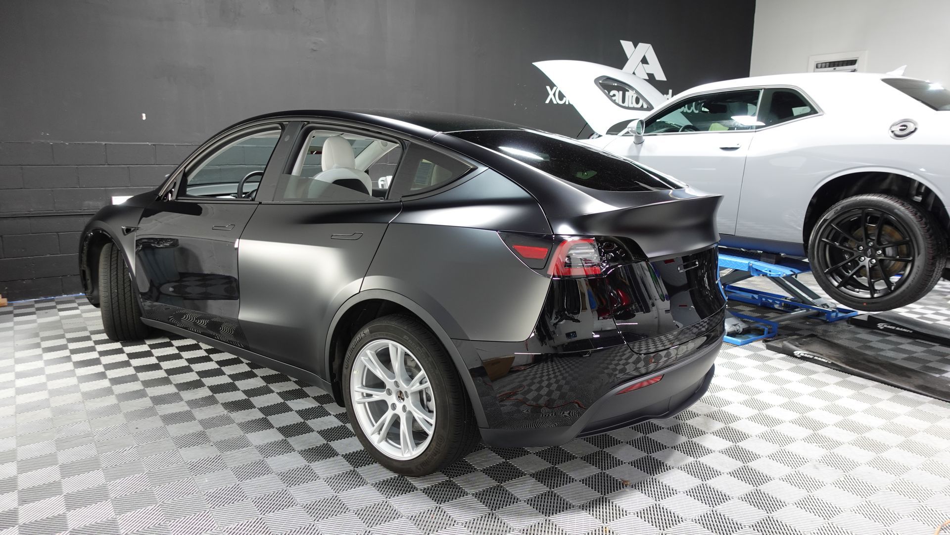 A black tesla model y is parked next to a white dodge challenger in a garage.