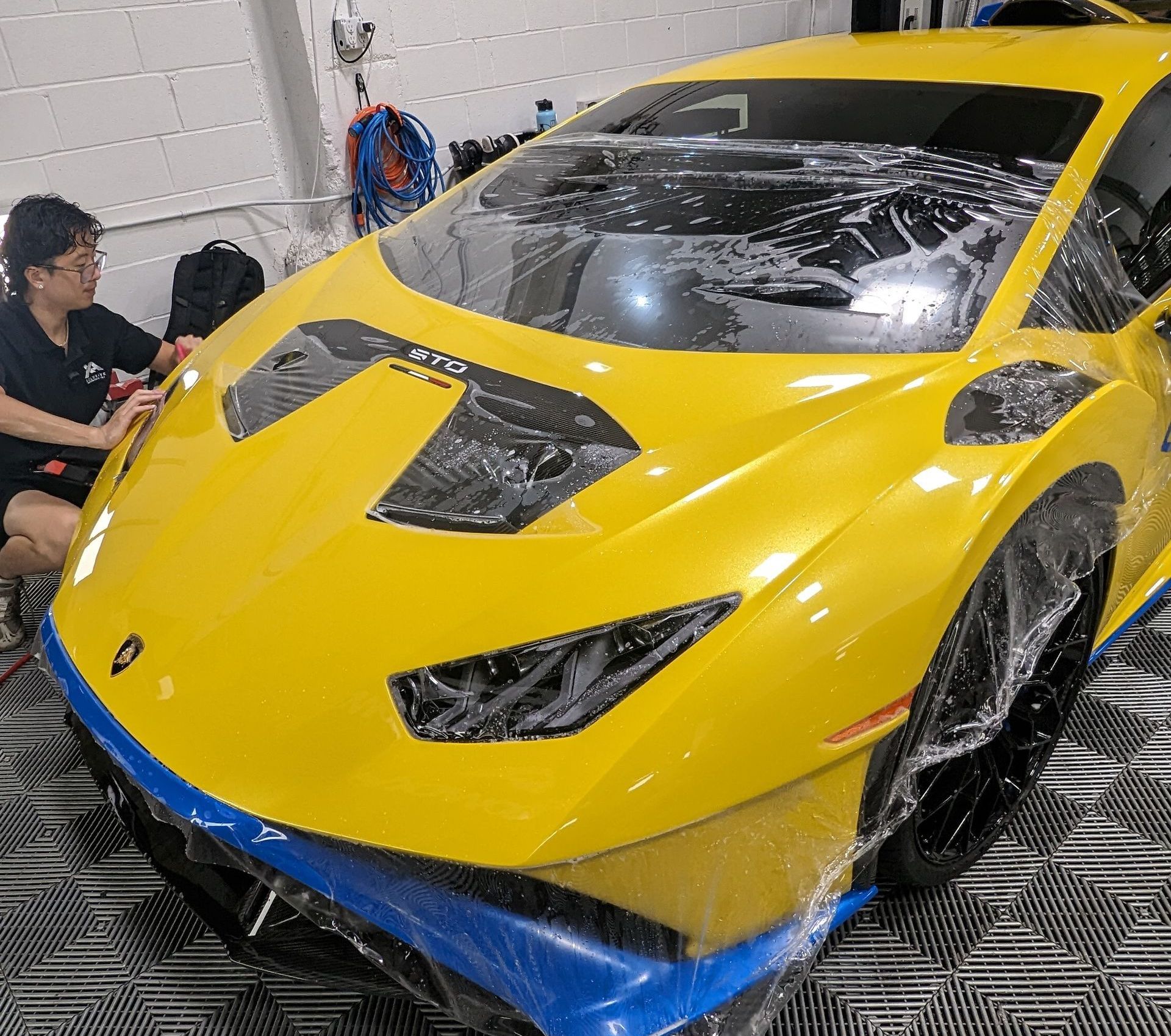 A man is sitting next to a yellow sports car wrapped in plastic.