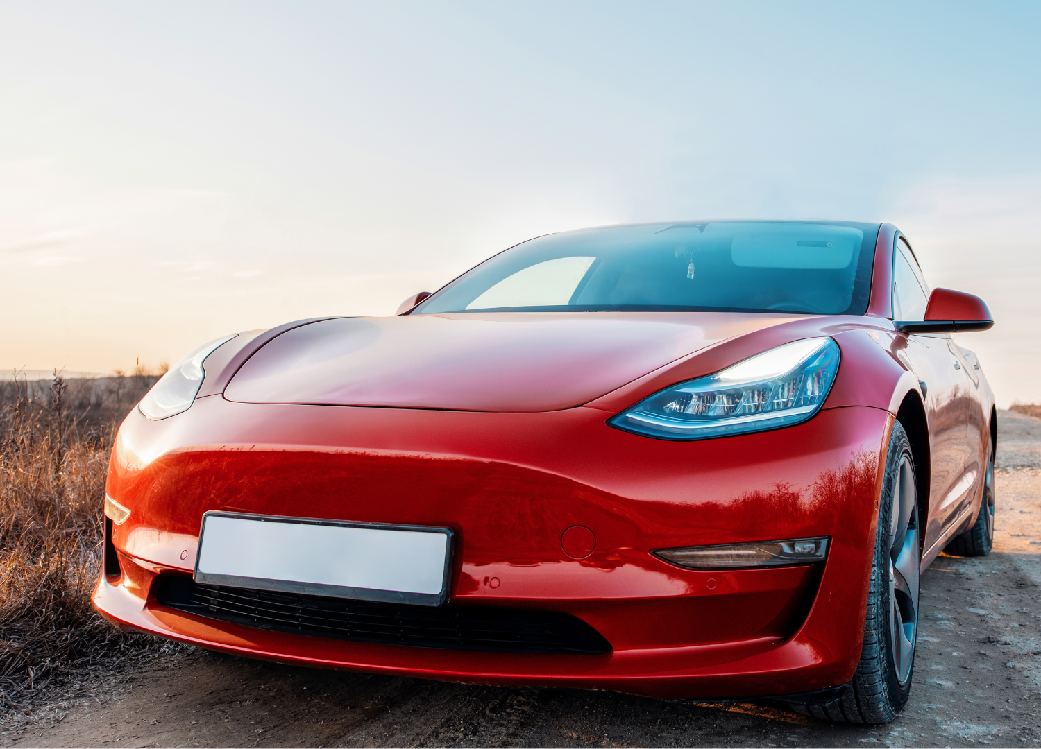 A red tesla model 3 is parked on the side of a dirt road.