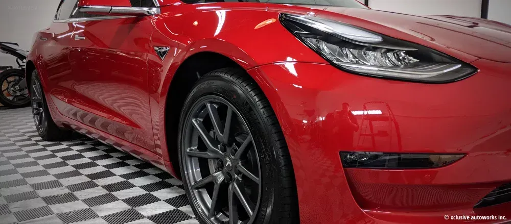 A red tesla model 3 is parked on a checkered floor in a garage.