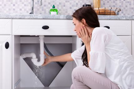 A woman is looking under a sink that is leaking water.