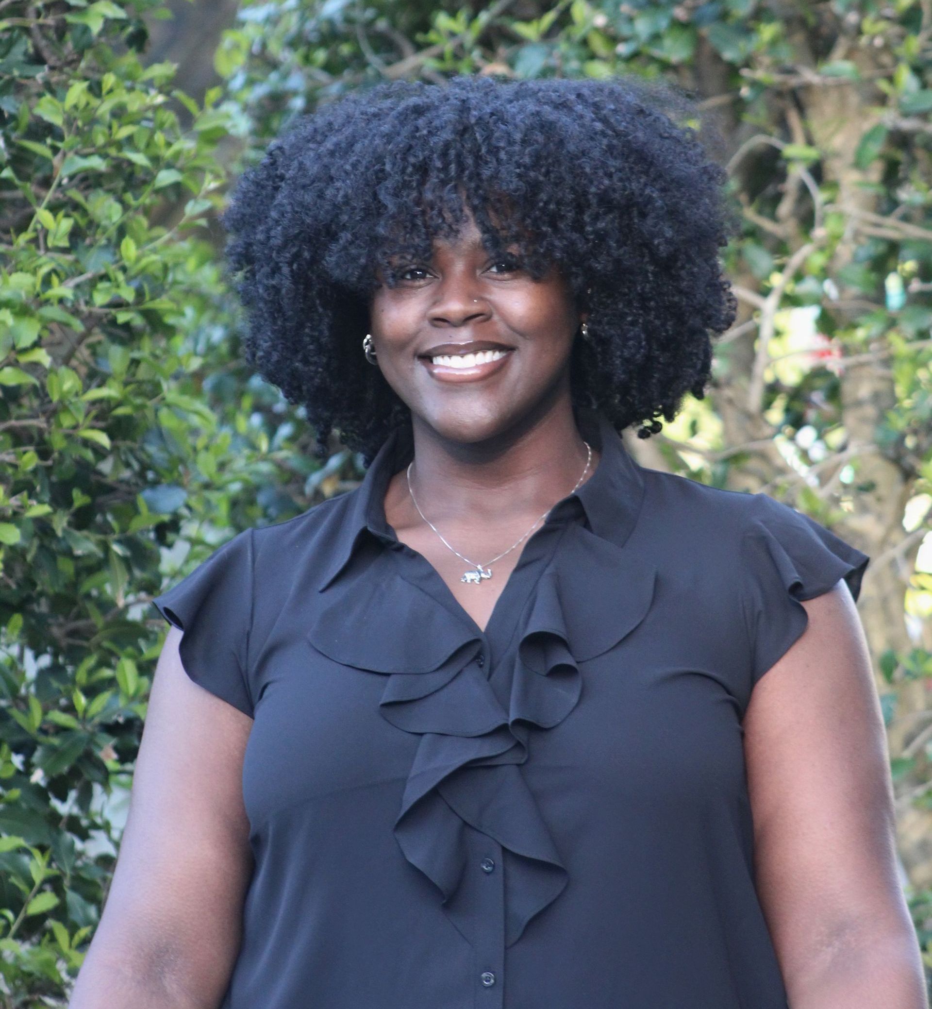 A smiling Black woman with large, dark hair poses outdoors. She wears a black blouse with ruffle detailing and a necklace.