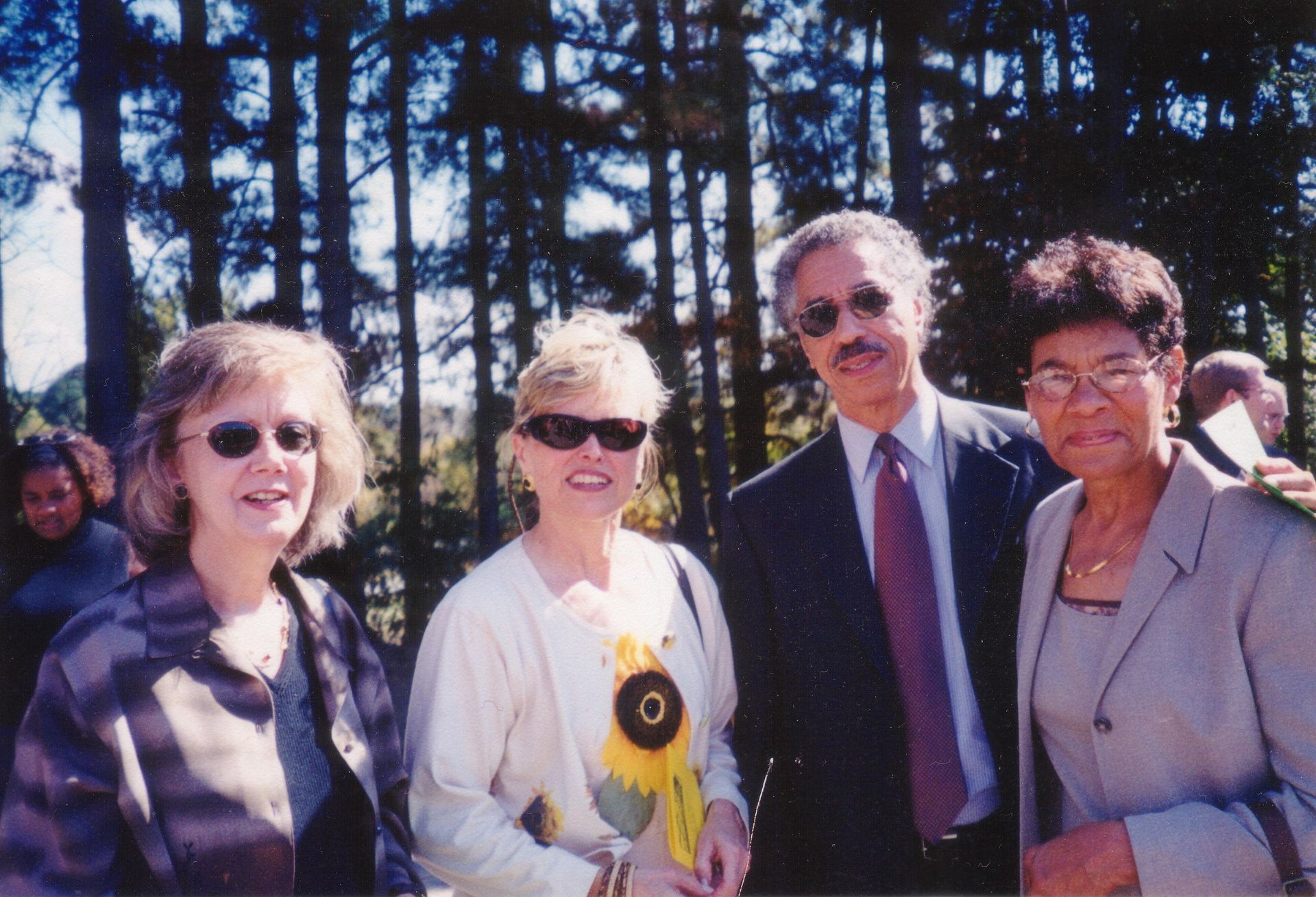 Four people smiling outdoors. Two women and a man are in the foreground; the other woman in the background has her arm around the man.