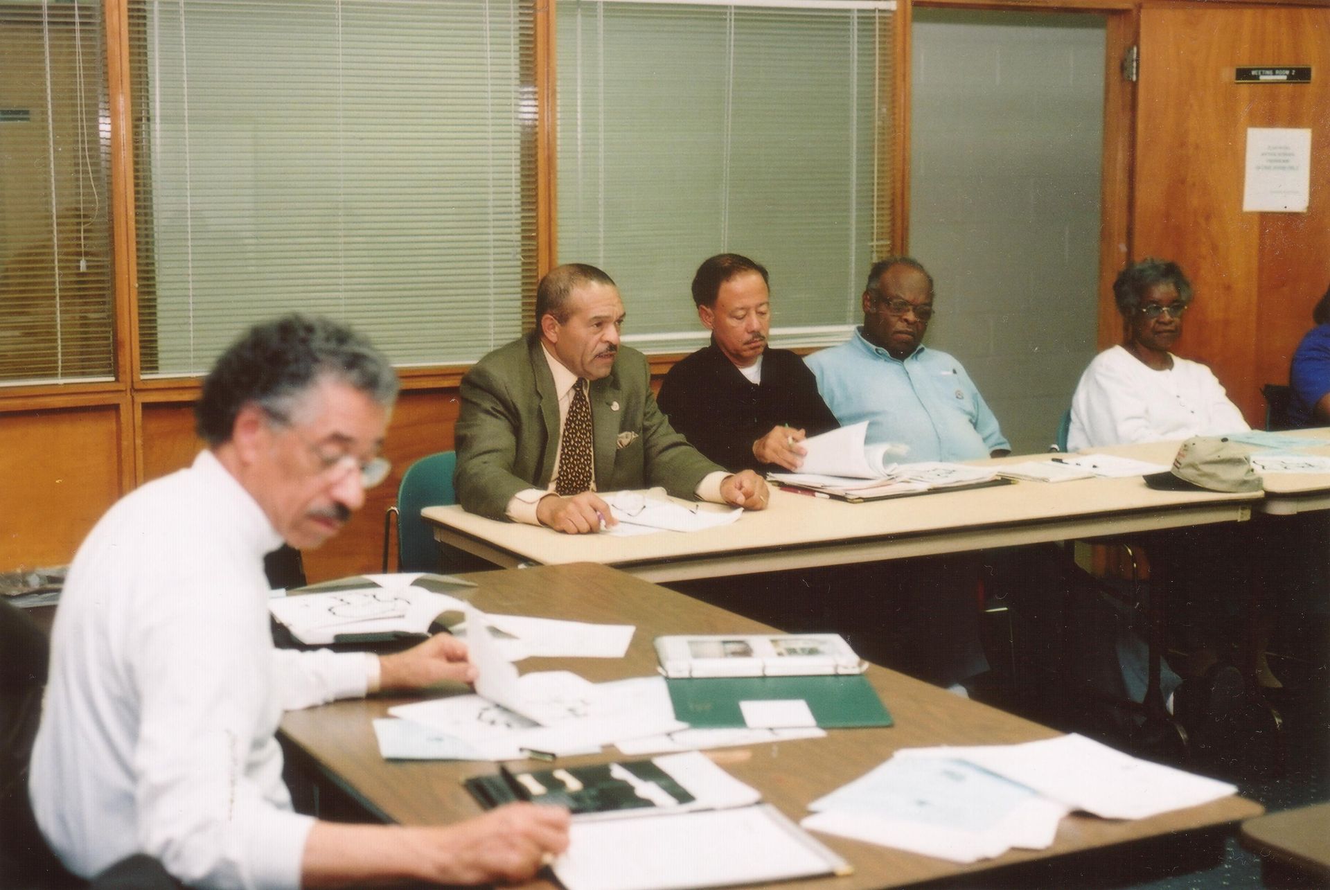 Five people seated at a table in a meeting, reviewing documents. They are in a brightly lit room with blinds on the windows.