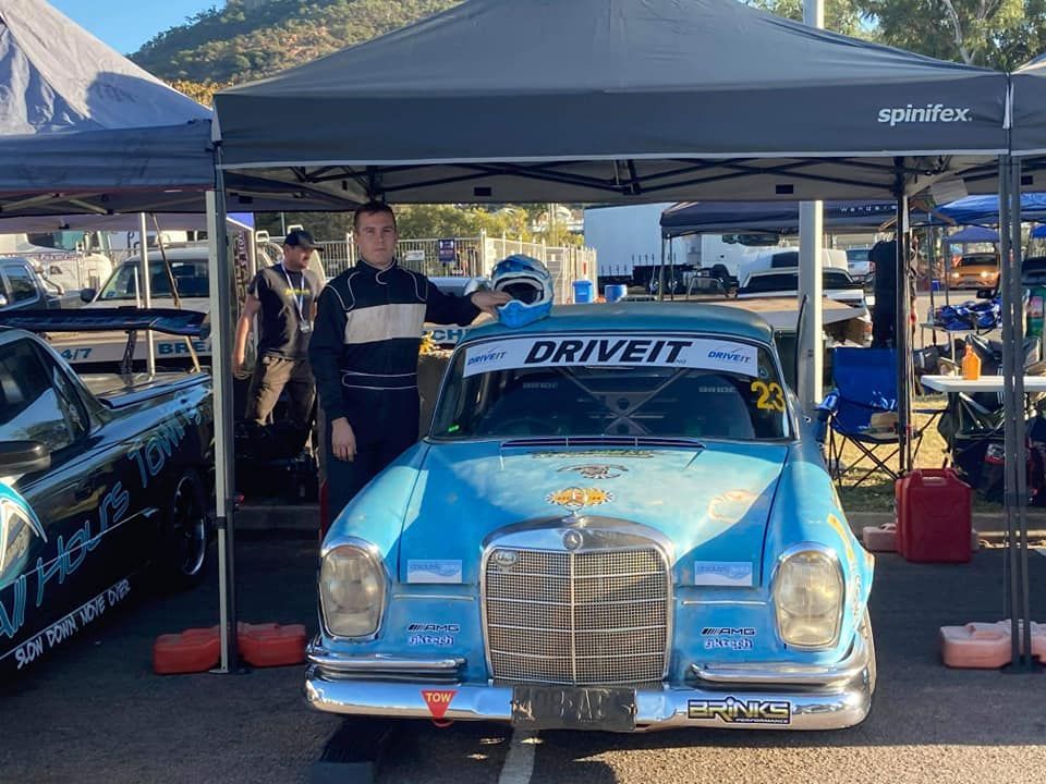 Race Car Driver in Uniform Poses Next to a Blue Vintage Mercedes Race Car Under a Tent. the Car Has 