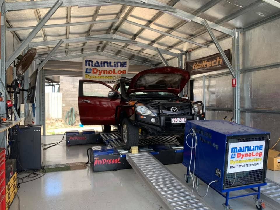 A Red Pickup Truck on a Dyno Inside a Garage. the Hood is Open, and a Technician is Likely Testing the Engine — Shannon's Auto Electrical in Garbutt, QLD