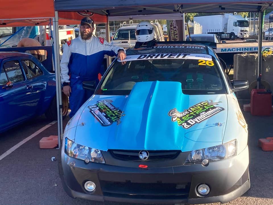 Man in Blue Race Suit Leaning on a Blue and White Race Car With a Large Hood Scoop. the Car is Under a Tent at a Race Track — Shannon's Auto Electrical in Garbutt, QLD