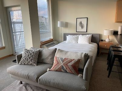 Bright bedroom with a bed, loveseat, nightstands, and large windows.