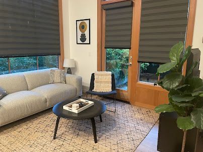 Cozy living room with gray sofa, round coffee table, armchair, and large windows with dark blinds.