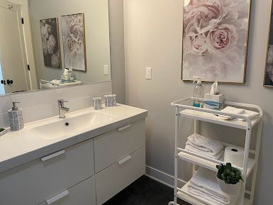 Bright bathroom vanity with mirror, sink, gray cabinets, wall art, and a small white shelving cart.