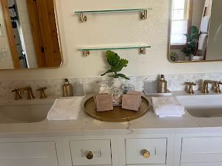 Double vanity bathroom with gold fixtures, white sinks, towels, and a tray with greenery centerpiece