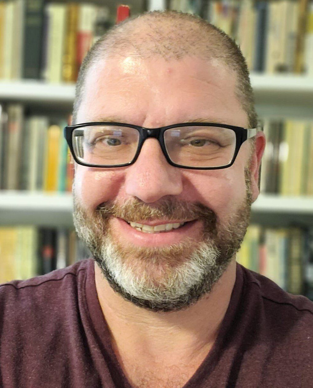 Man with glasses and beard smiles, wearing a maroon shirt, bookshelves in background.