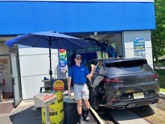 An attendant in a blue polo and light shorts stands by a car at a wash tunnel entrance under a blue umbrella.