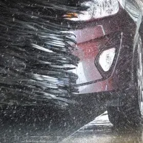 A close-up view of a car undergoing a mechanical car wash with rotating black brushes scrubbing the bumper.