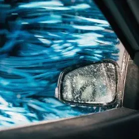 A car side mirror obscured by water droplets and blurry blue automatic car wash brushes outside a window.