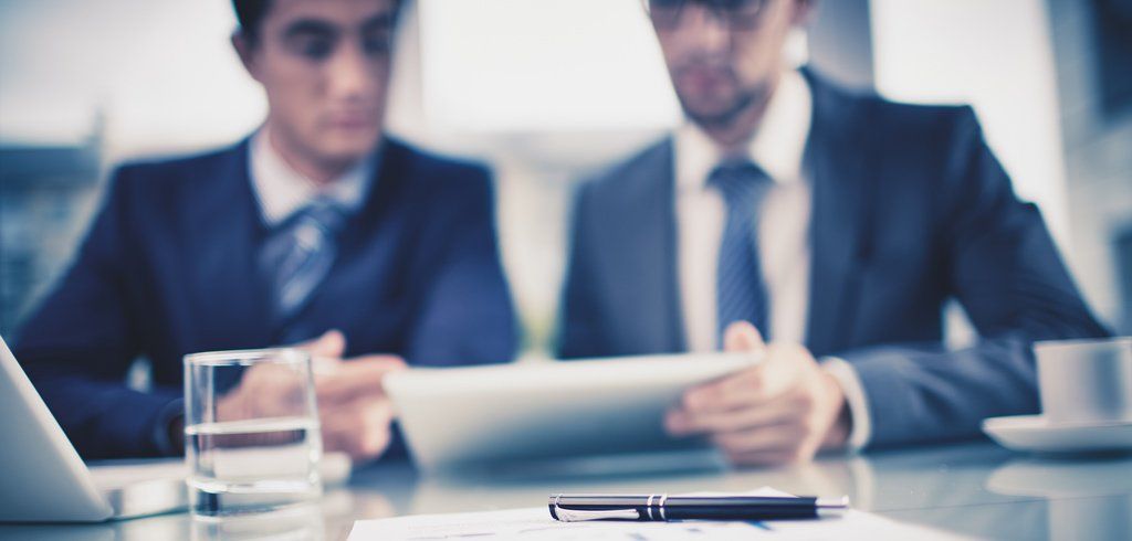 Two men in suits and ties are sitting at a table looking at a tablet.