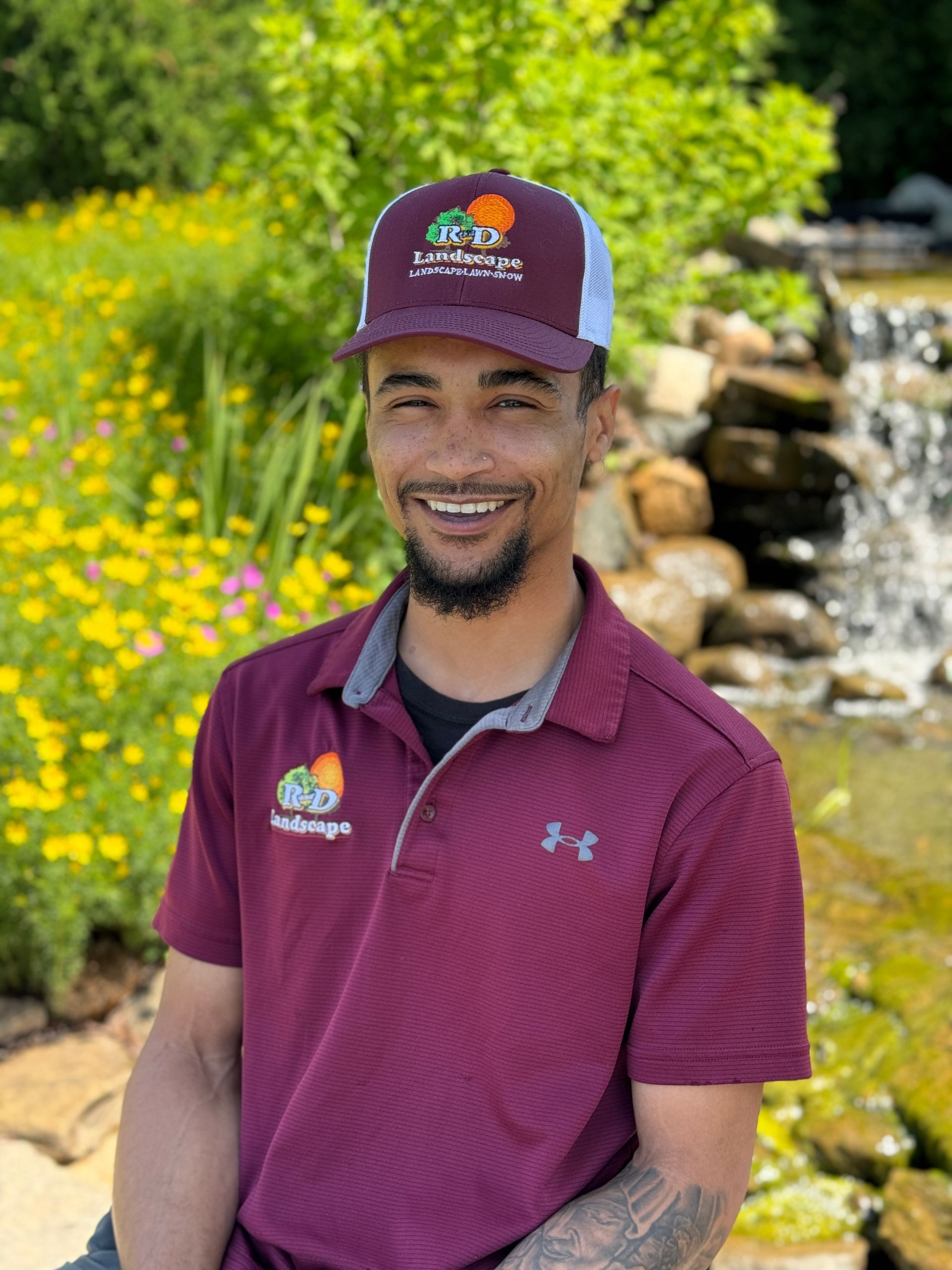 A man wearing a hat and a maroon shirt is standing in front of a waterfall.