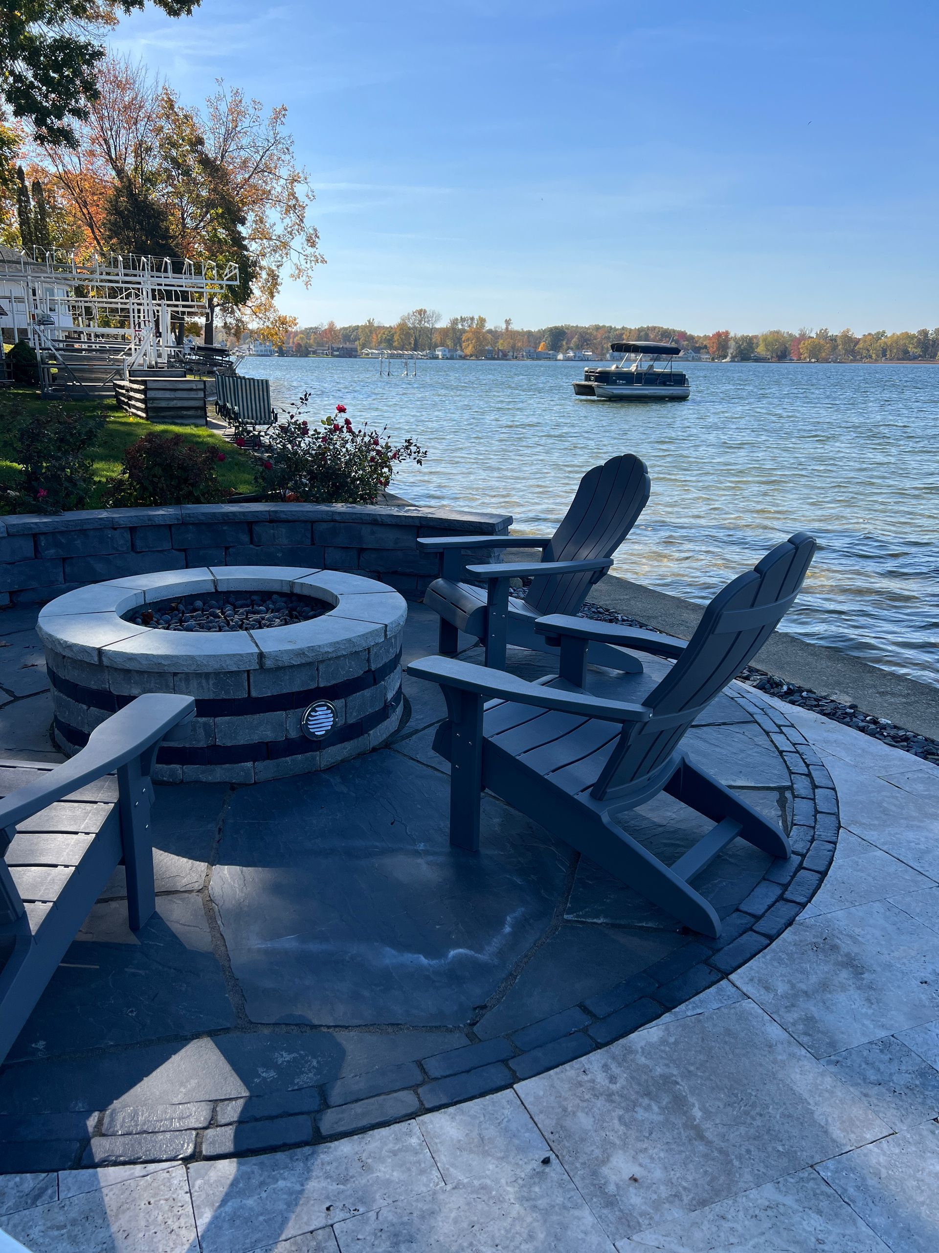 A fire pit is surrounded by chairs on a patio next to a lake.