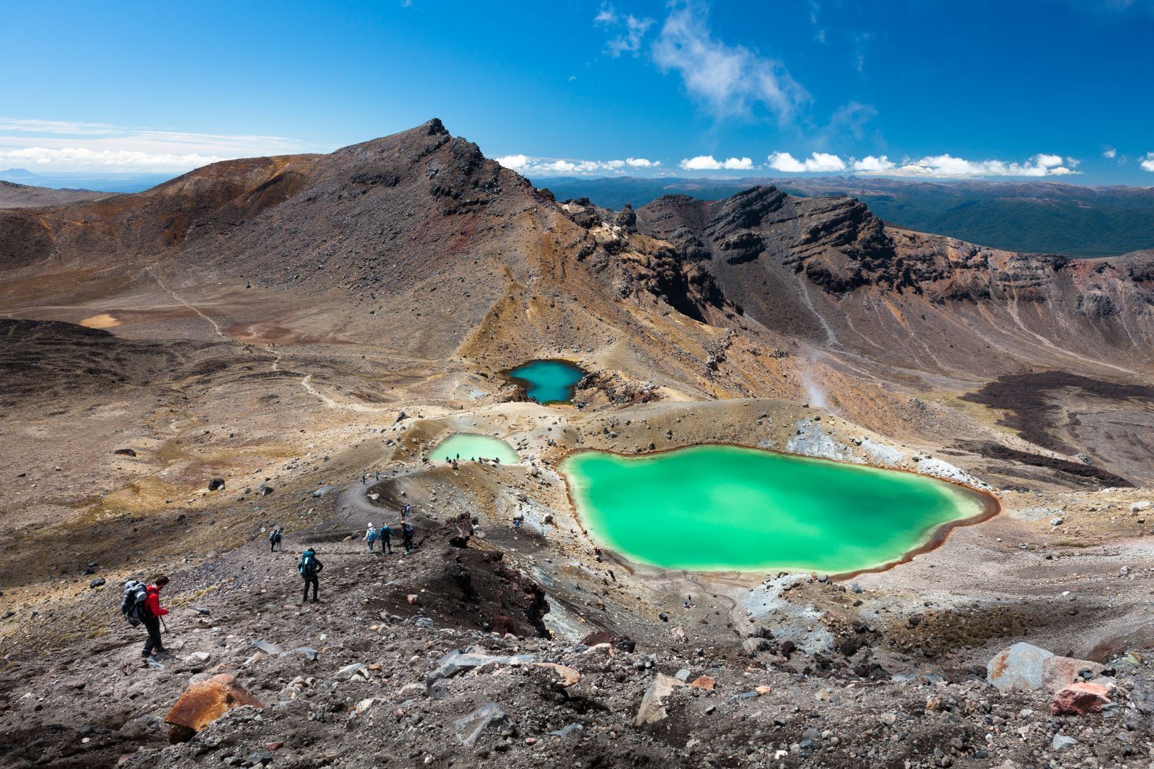 Hikers overlooking bright green crater lakes in a rocky volcanic landscape under a blue sky