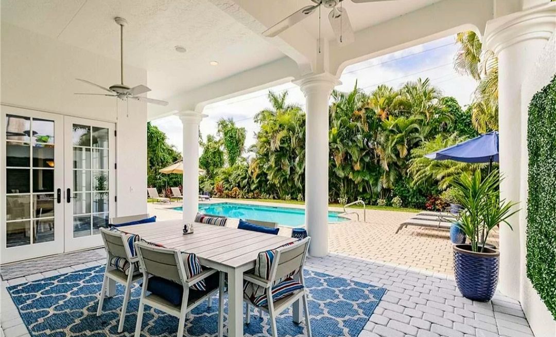 Covered patio with white dining set, blue rug, pool, and lush greenery beyond the columns