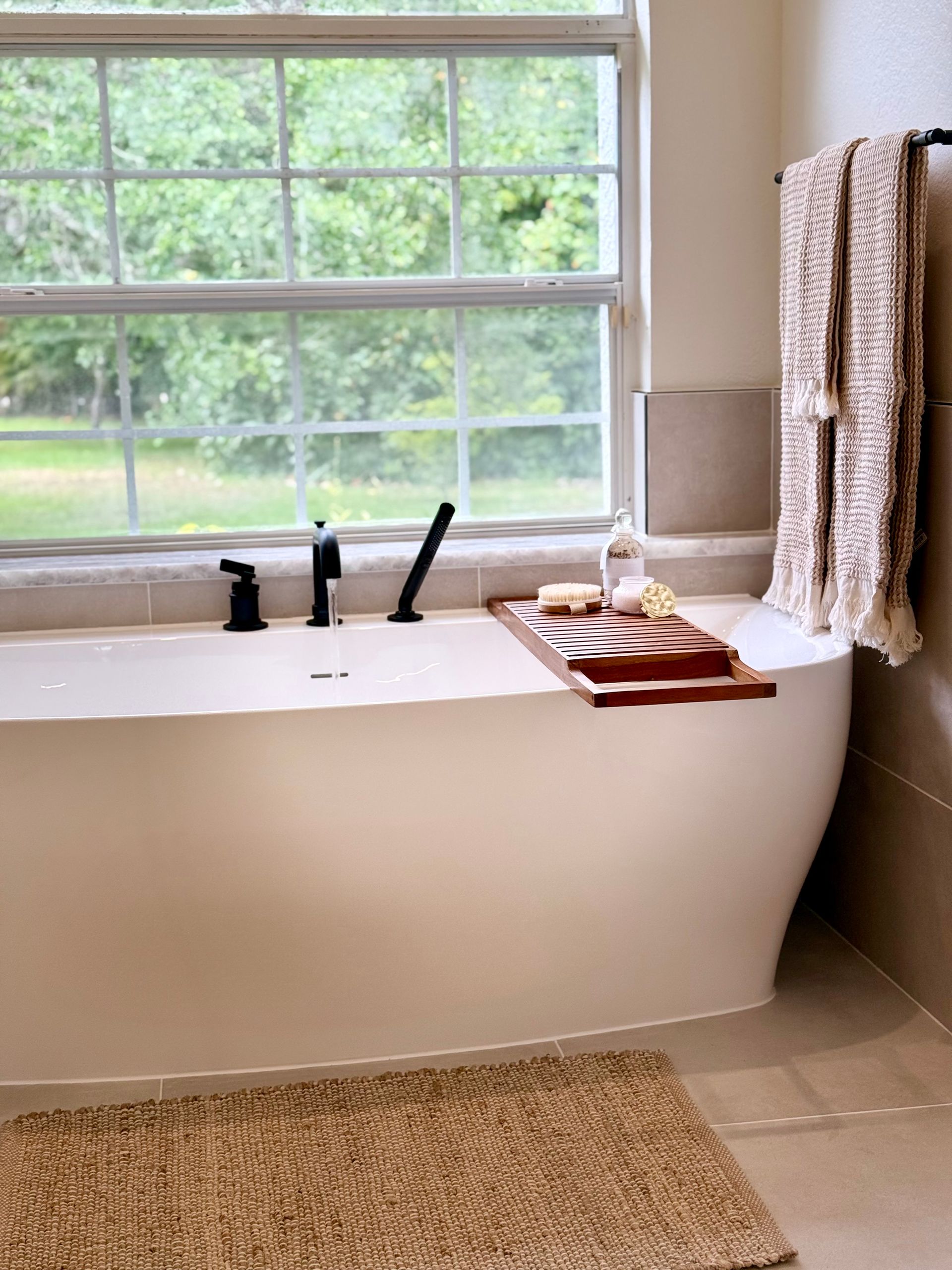 Modern bathroom with a freestanding tub, tray, towels, and a large window overlooking greenery