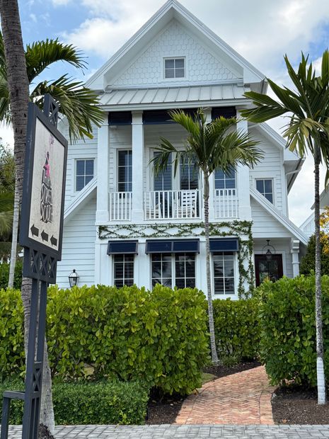 White two-story house with balconies, black shutters, and a brick path behind hedges and palm trees