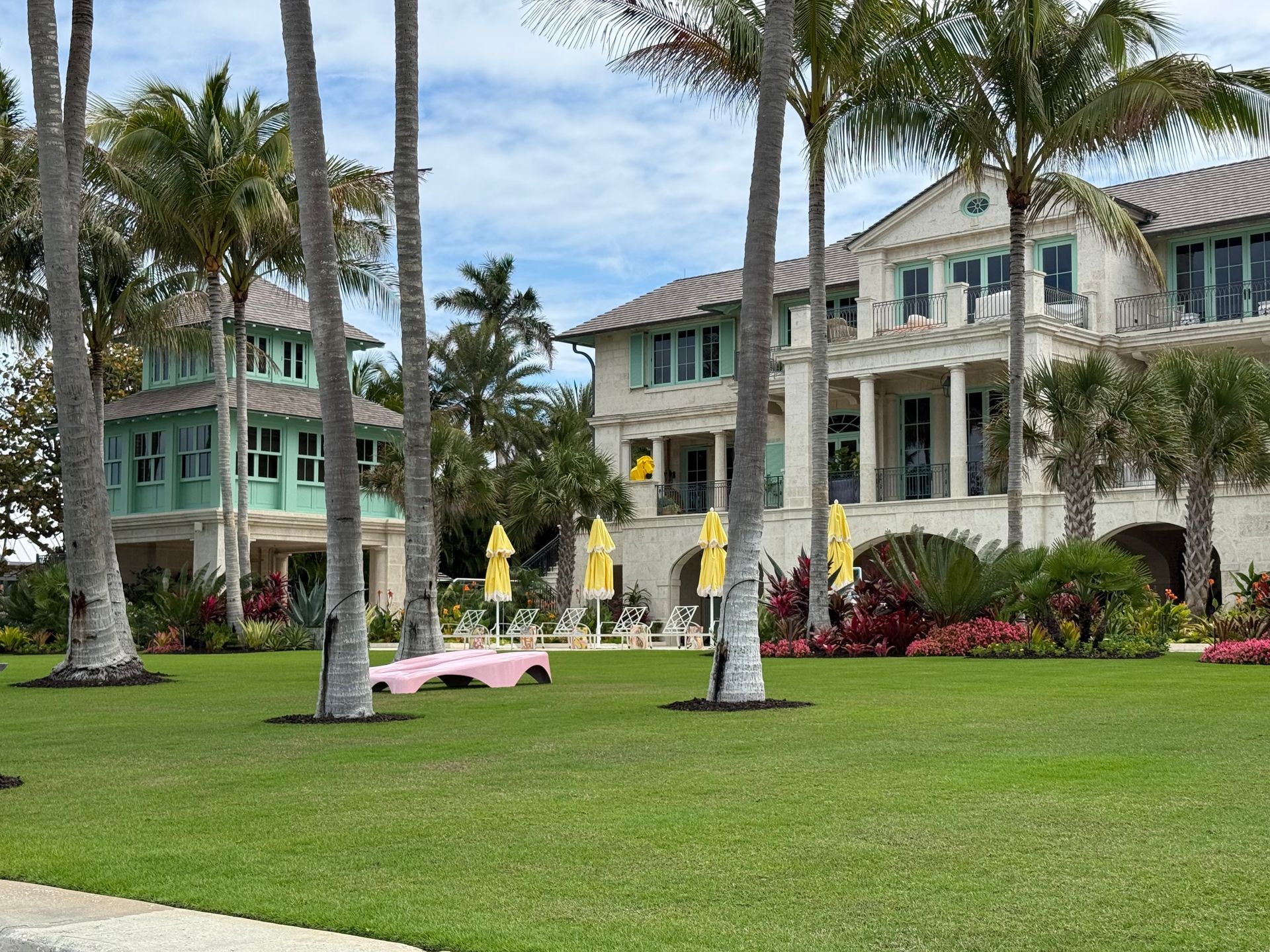 Lawn with palm trees, pink lounge chairs, and pastel resort buildings under a partly cloudy sky