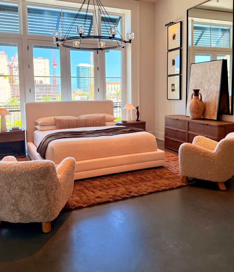Bright bedroom with a white bed, two fuzzy chairs, wicker dresser, and large windows with city view.