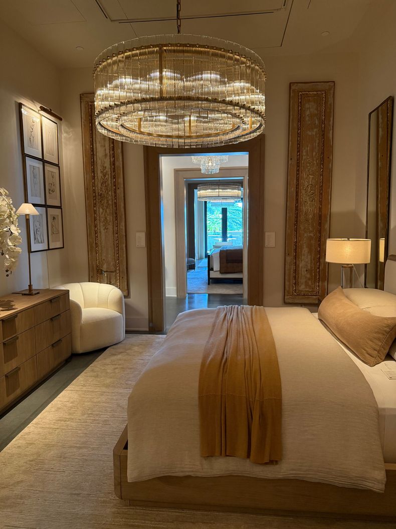 Elegant bedroom with a large chandelier, beige decor, twin beds, and a bright doorway to a bathroom.