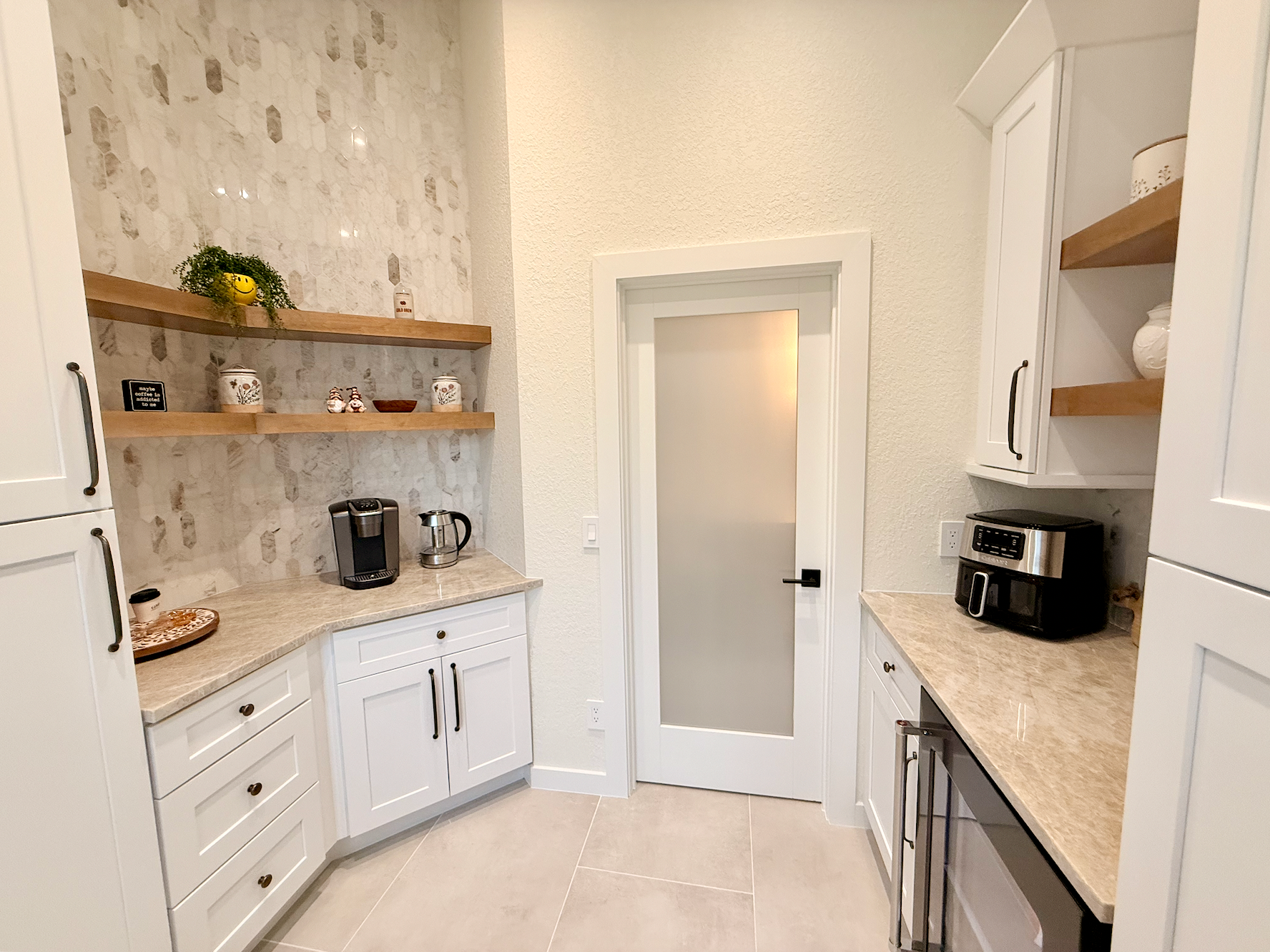 Bright white kitchen with marble counters, open shelves, a coffee maker, and a frosted glass door.