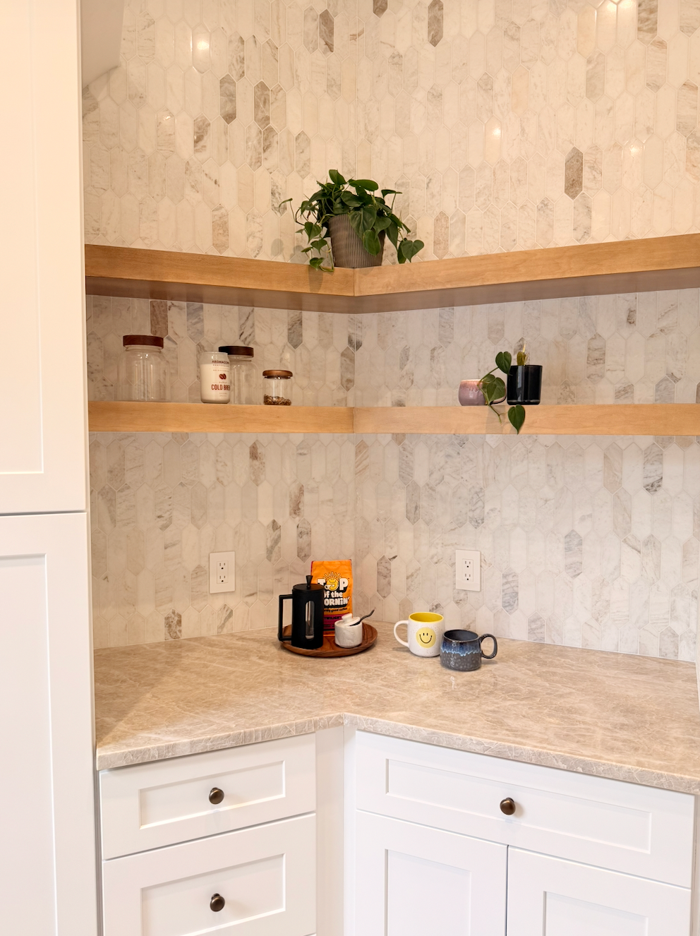Corner kitchen countertop with white cabinets, marble-look backsplash, and shelves holding small plants and mugs