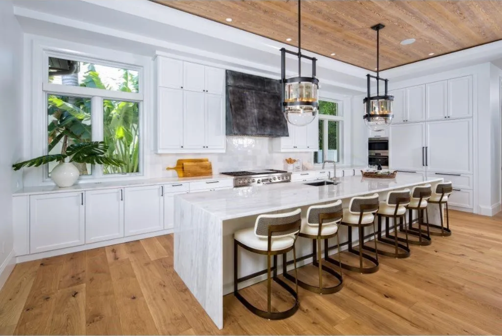 Bright white kitchen with island, bar stools, hanging lights, wood floor, and large windows