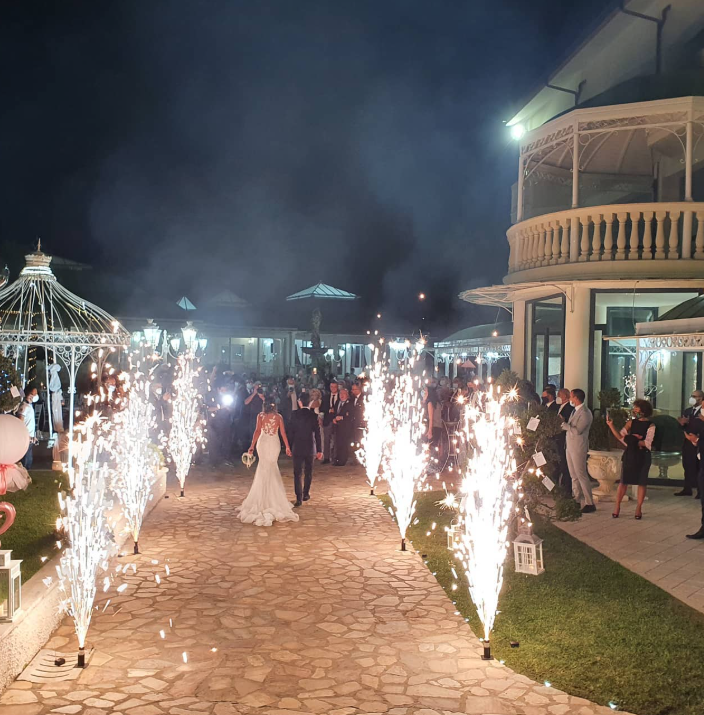 Gli sposi percorrono un sentiero di pietra fiancheggiato da fuochi d'artificio durante un ricevimento di nozze notturno. Gli ospiti guardano.